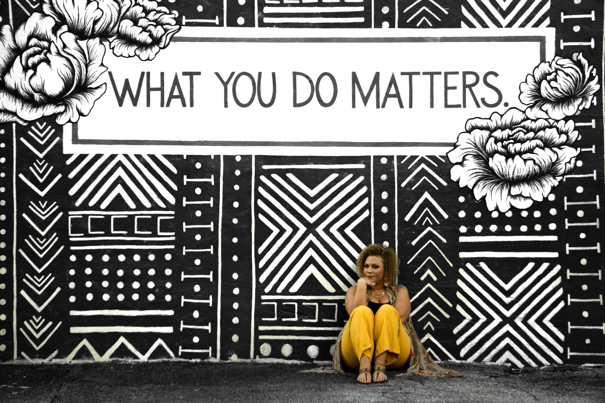 Künstlerisches Wandbild mit floralen und geometrischen Mustern in Schwarz-Weiss und dem zentralen Schriftzug „What you do matters“, vor dem eine Frau mit gelber Hose sitzt.