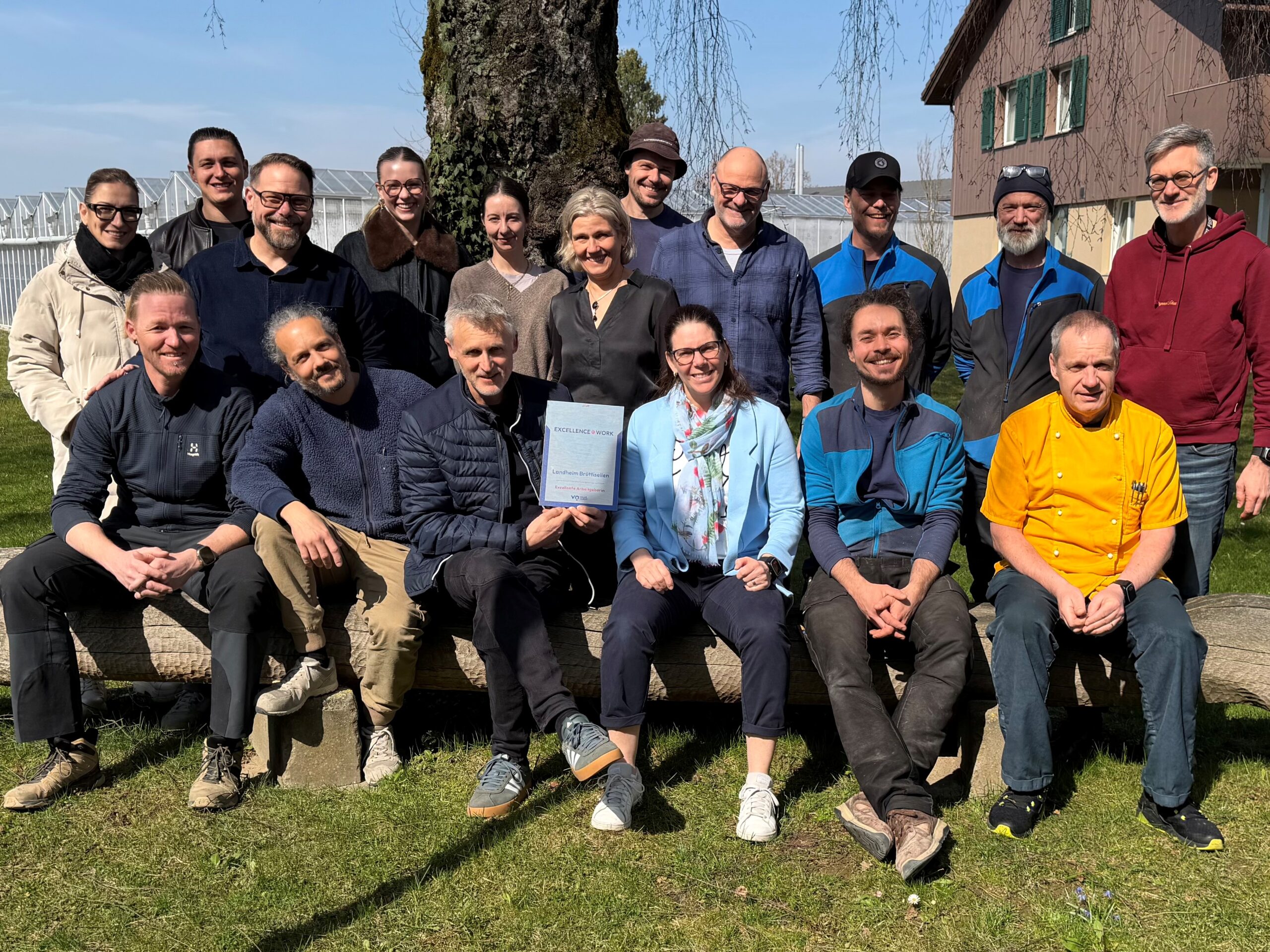 Gruppenfoto des Teams vom Landheim Brüttisellen im Freien bei sonnigem Wetter. In der Mitte wird ein Excellence@work Award gehalten, der die Auszeichnung als Top-Arbeitgeber bestätigt.