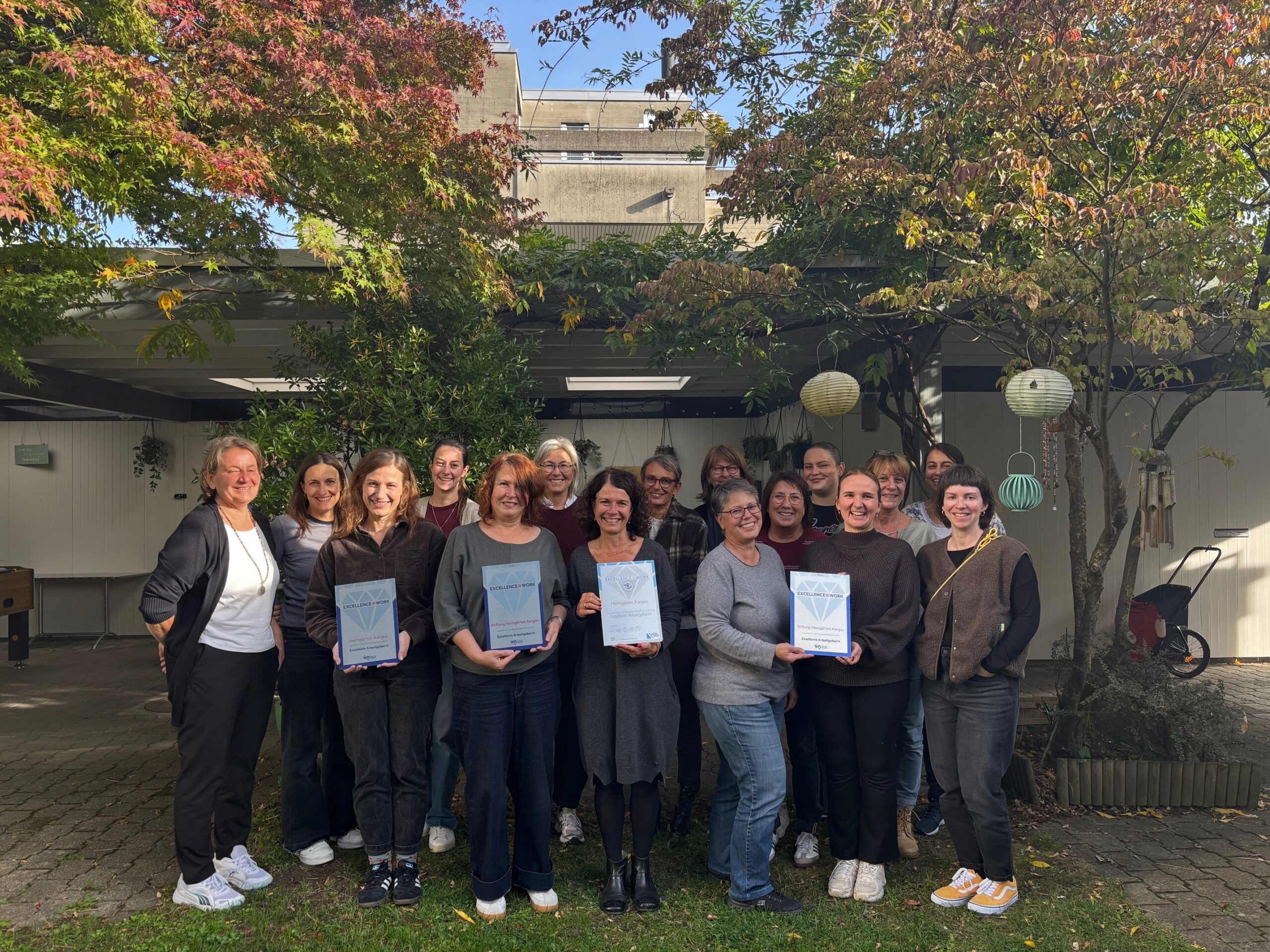 A group photo of the Heimgärten Aargau team outdoors on a sunny day. They are holding four Excellence@work Awards: the award for Top Employer.